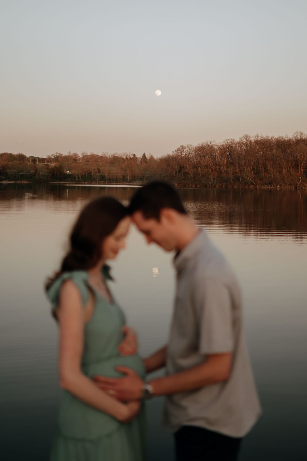 Couple touching foreheads over pregnant belly at dusk with full moon reflecting on calm lake.