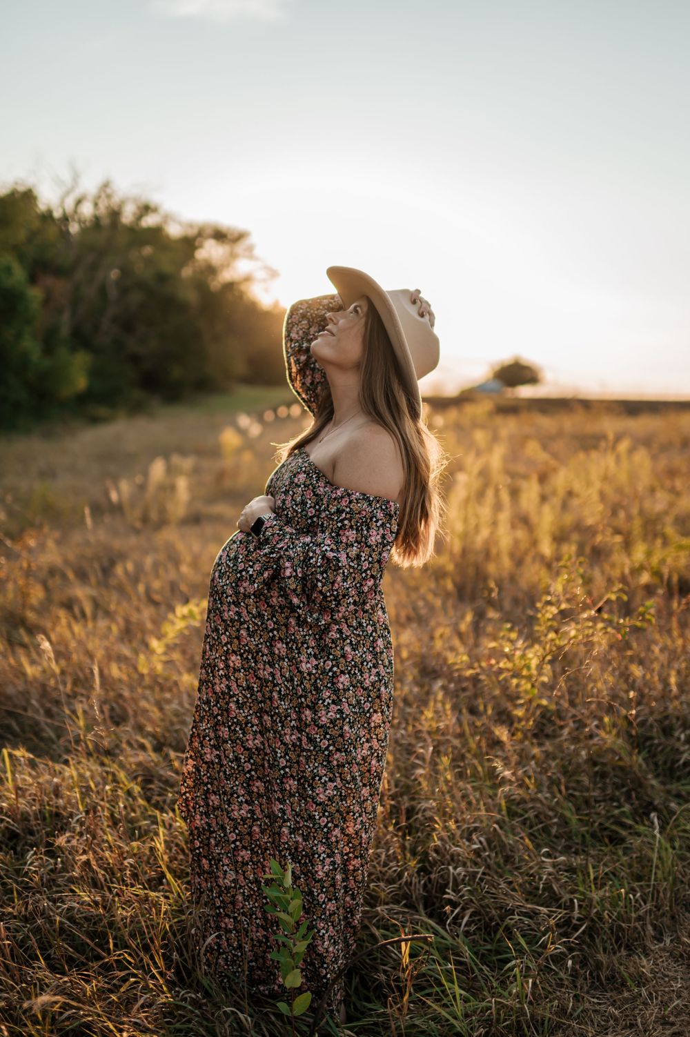 Pregnant woman in floral dress and cowboy hat gazing upward in golden sunlit field at sunset.