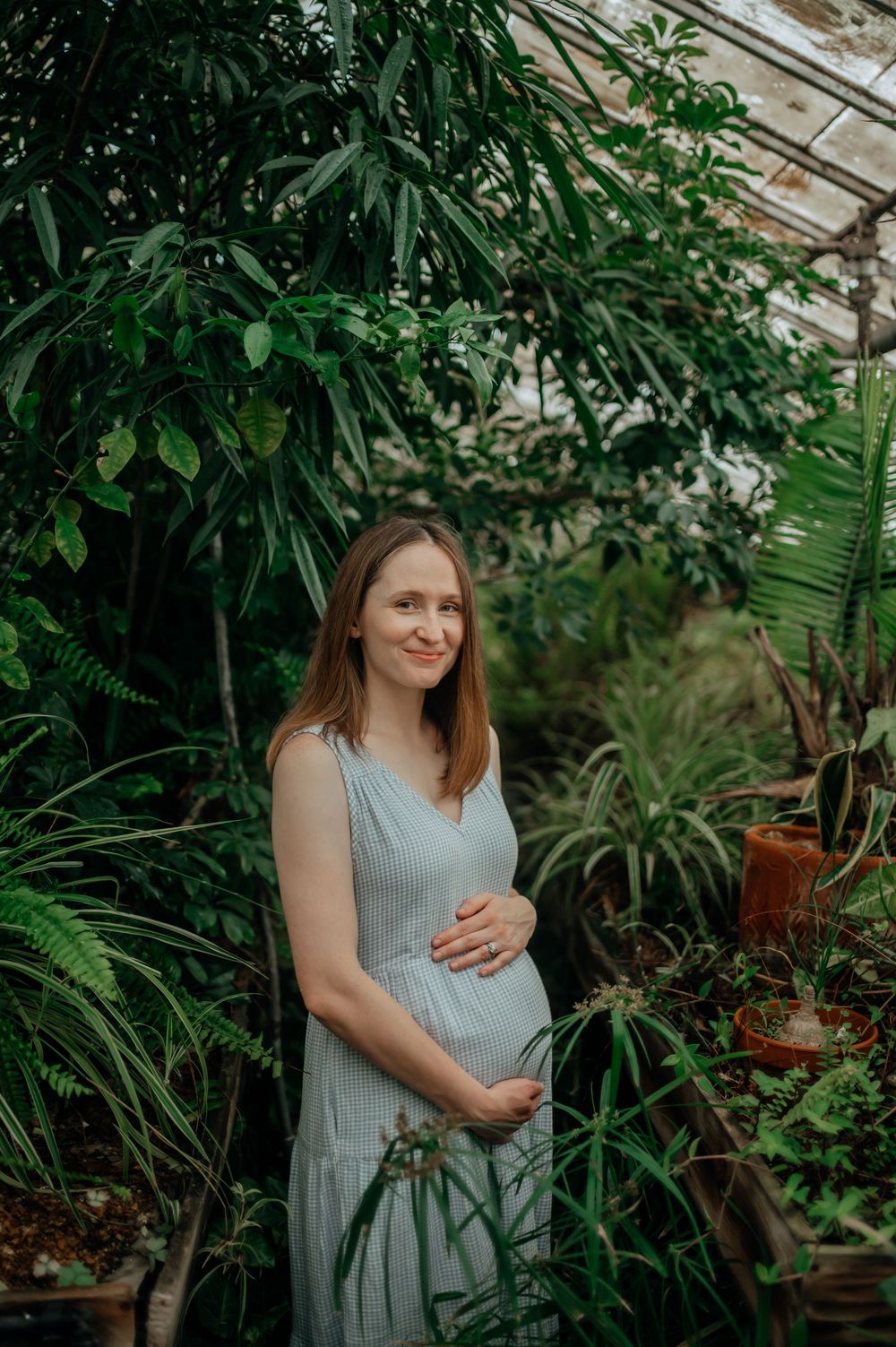 Pregnant woman in light blue dress holds baby bump in lush green greenhouse.