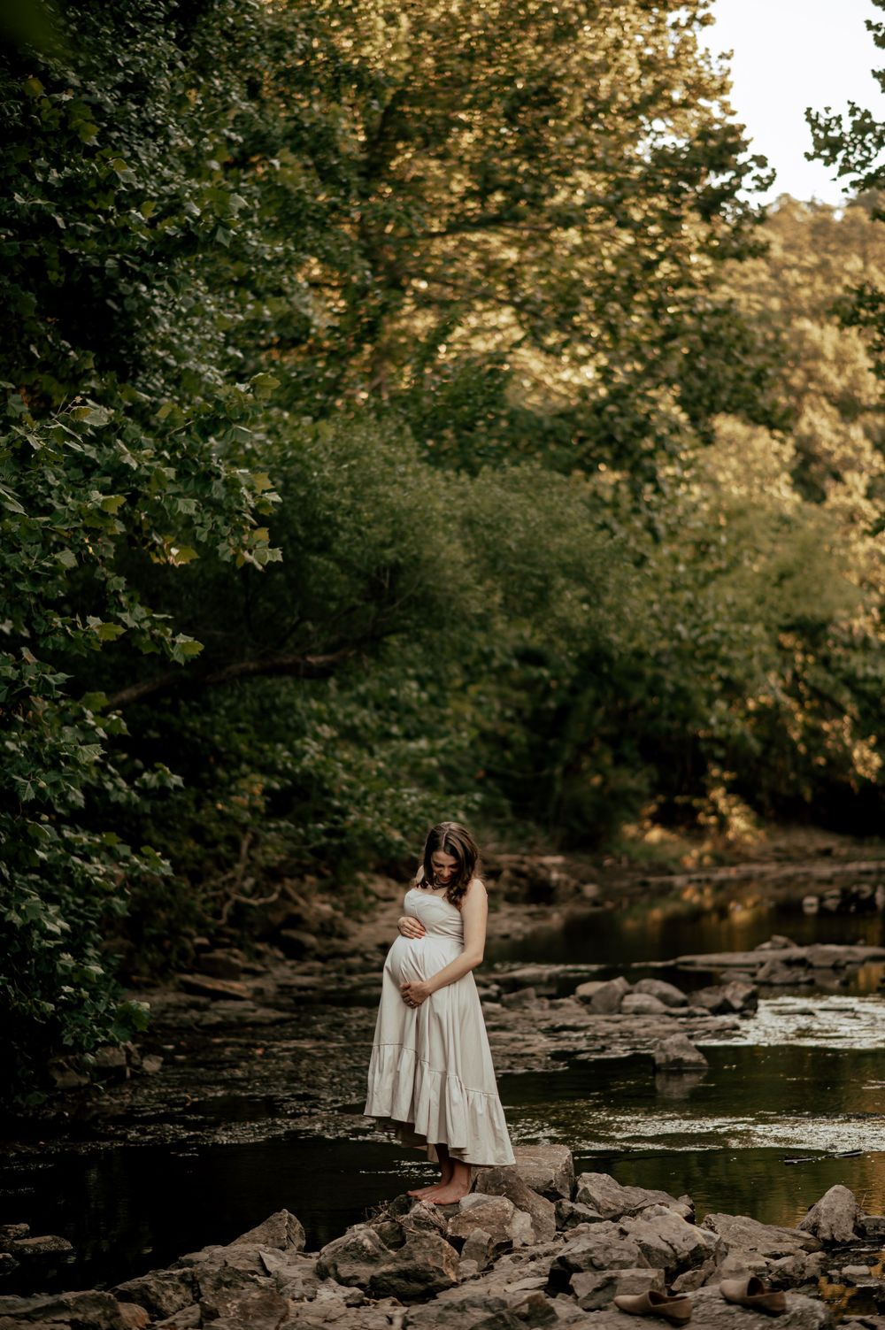 Pregnant woman in white dress cradling belly while standing on rocks beside a serene creek.