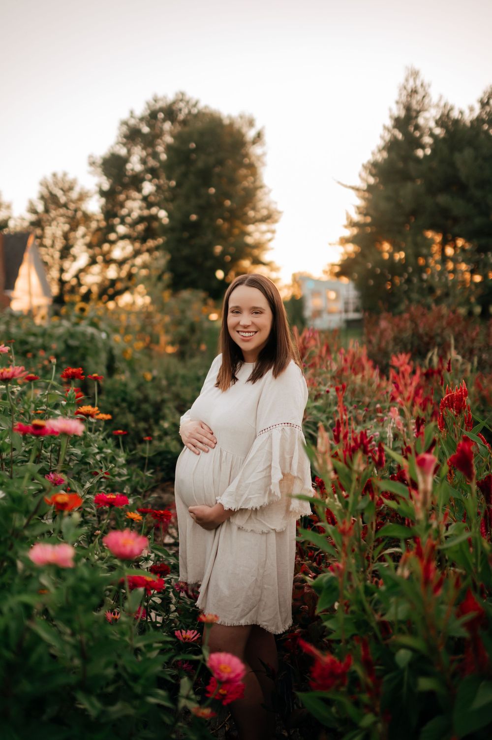 Smiling pregnant woman in white dress holds baby bump in colorful flower garden at sunset.