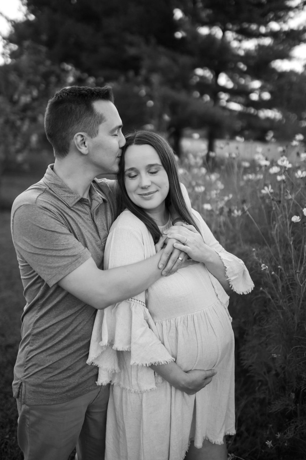 Black and white photo of man kissing pregnant womans temple while embracing in flower field.