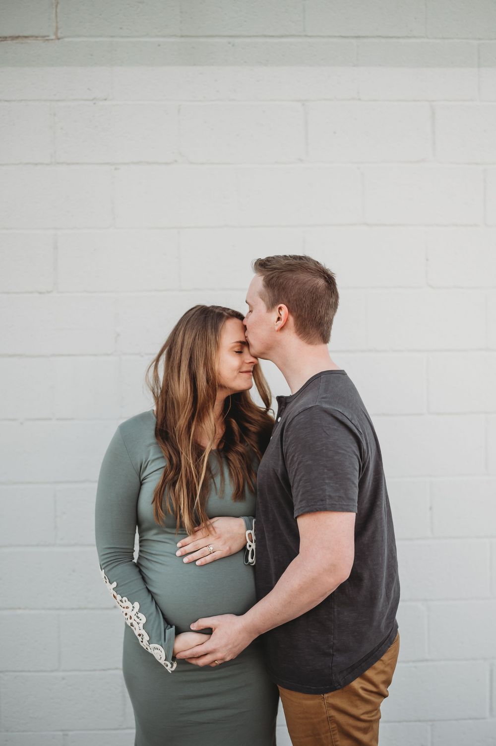 Pregnant woman in sage green dress embraced by partner kissing her forehead against white brick wall.