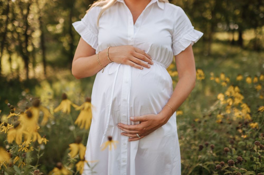 Close-up of pregnant woman in white dress cradling baby bump among yellow wildflowers.
