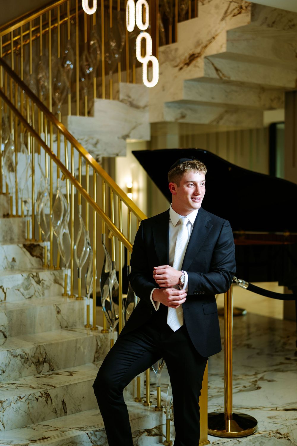 A groom in a black tuxedo leans against an elegant gold staircase railing in an ornate venue.