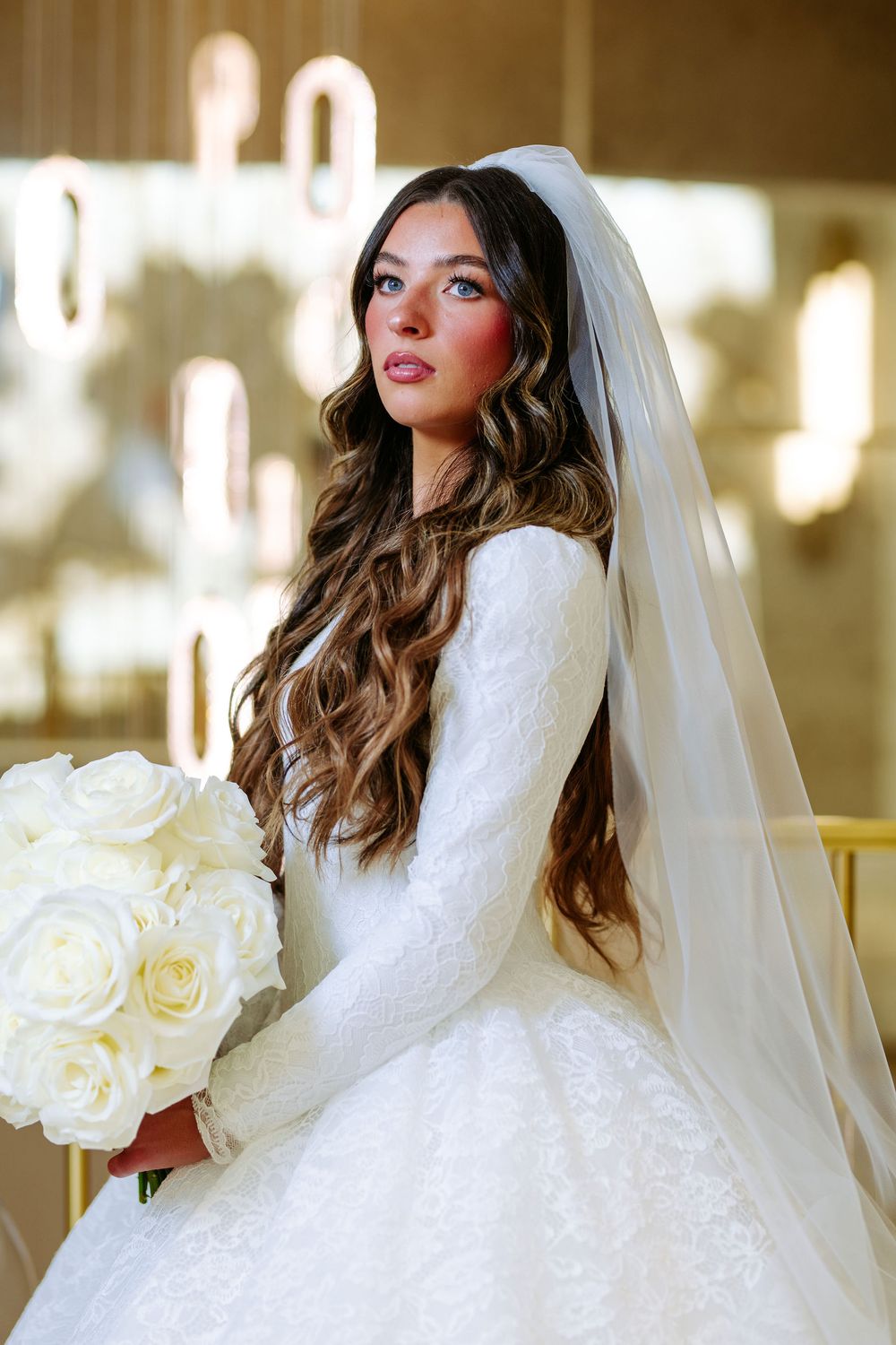 Bride in a white ballgown wedding dress with lace details holds a bouquet of white roses in an elegant indoor setting.