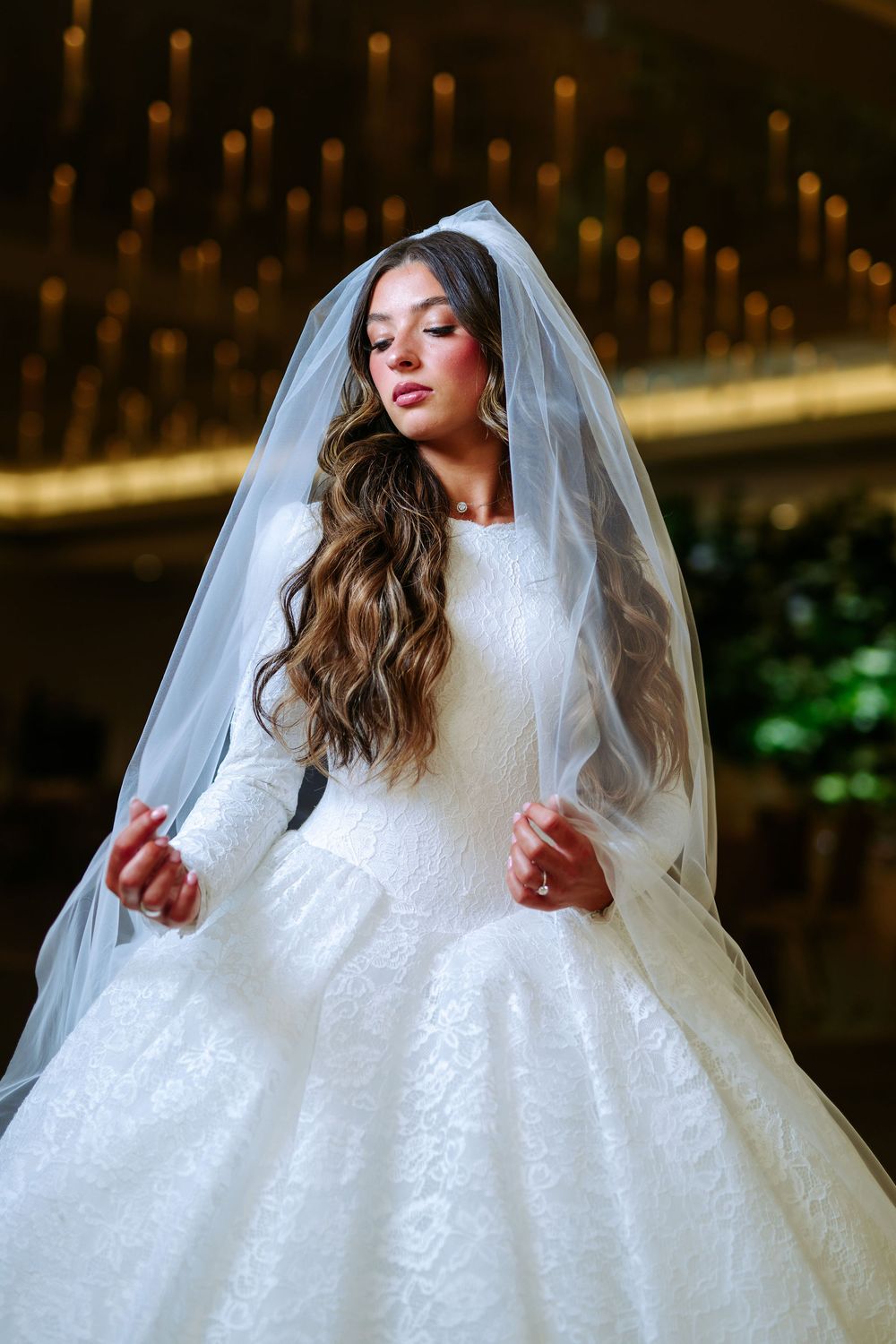 A bride in a white lace wedding dress and veil poses elegantly against a bokeh light background.