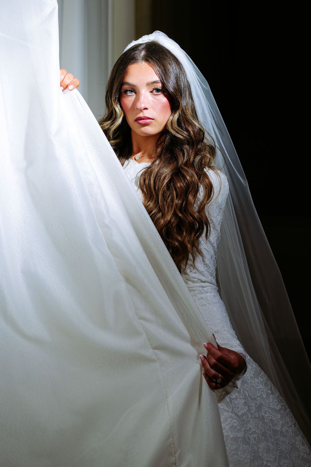 Bride in white lace wedding dress with flowing veil poses against dramatic lighting.