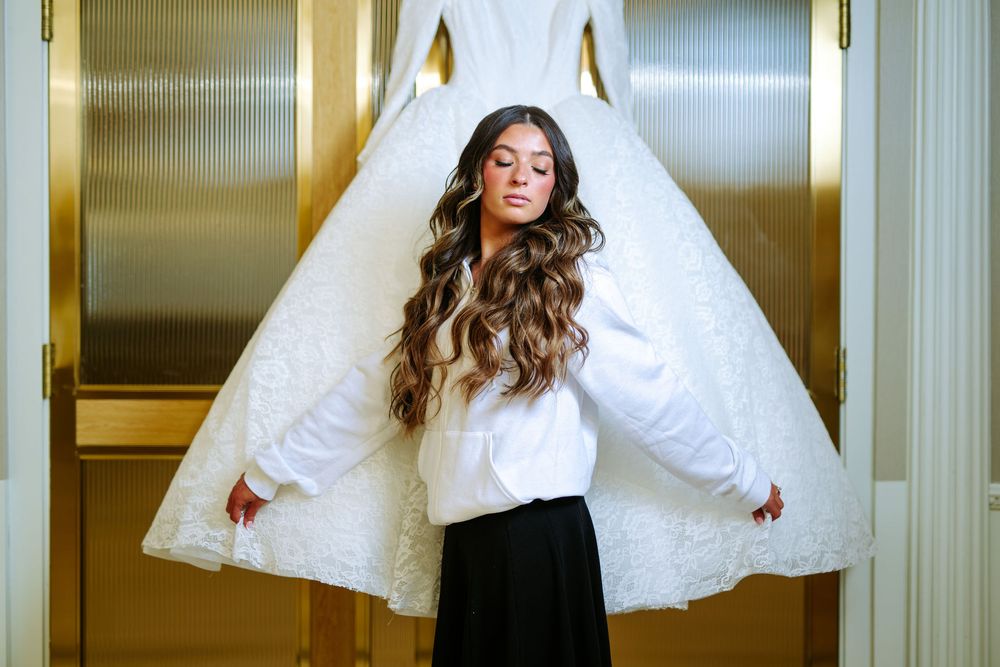 A bride in a black top playfully holds up her white tulle wedding dress skirt in an elegant hallway setting.