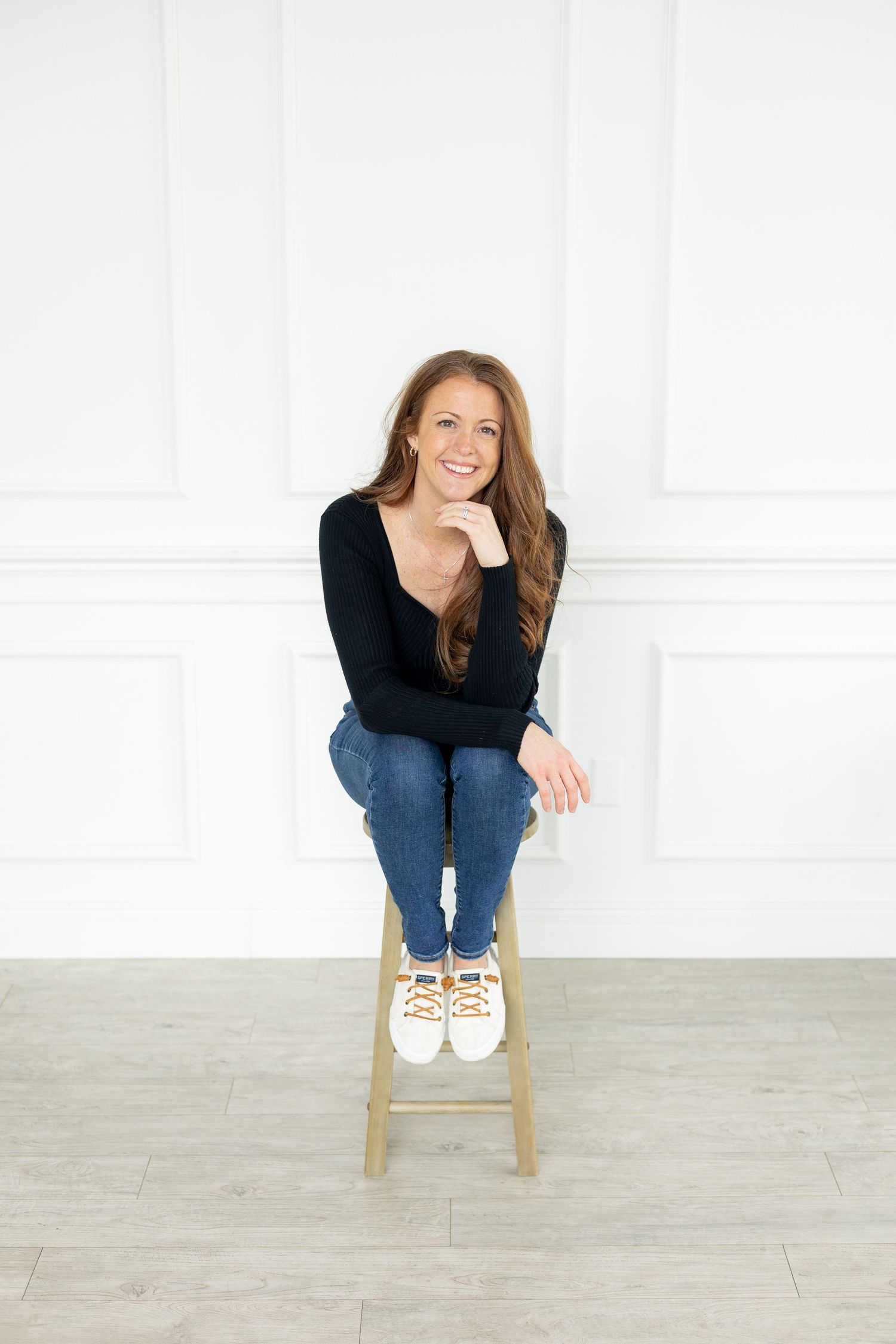 A person in a black sweater and jeans sits casually on a wooden stool against a white wall.