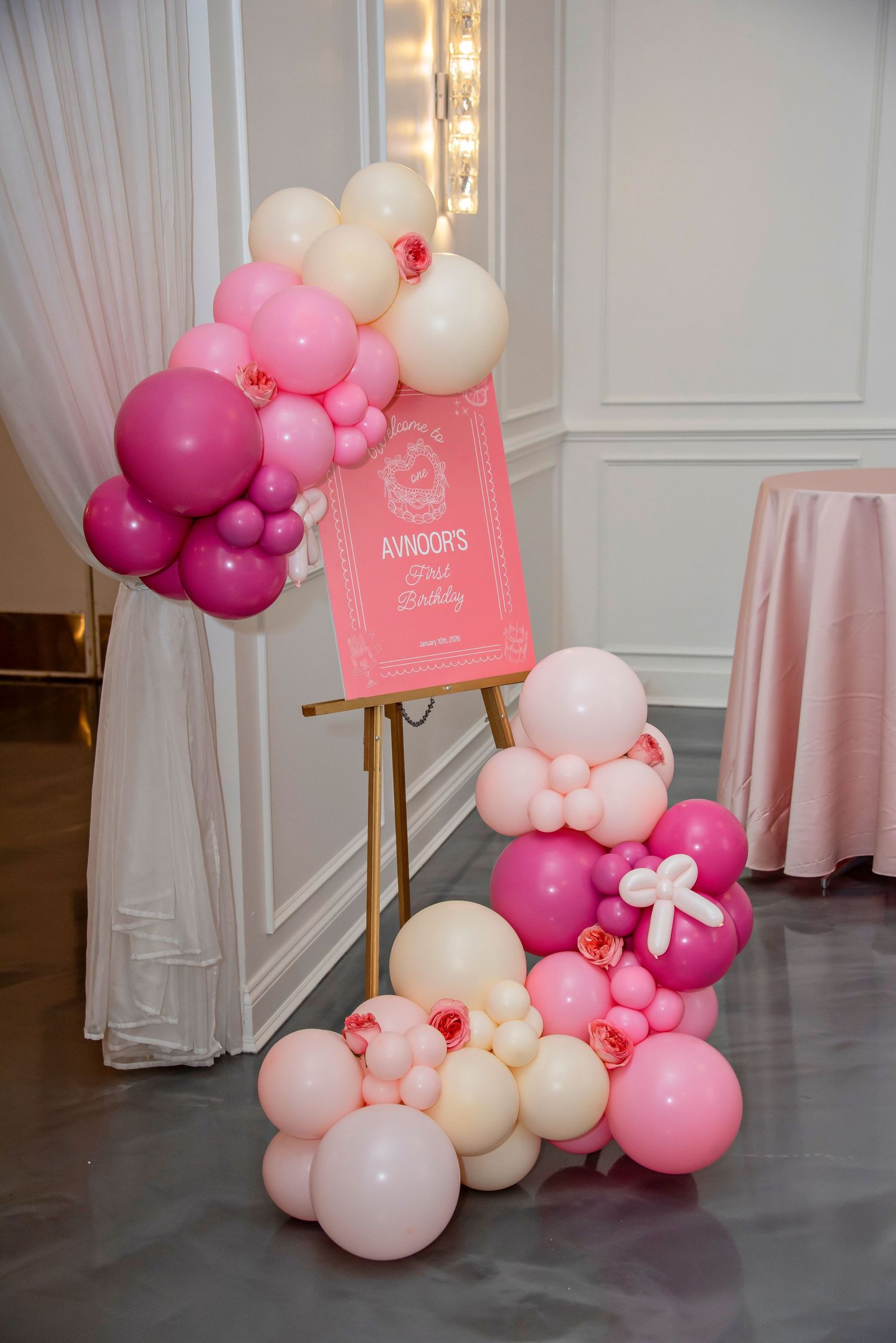 Pink and white balloon arch decoration with a pink sign on a gold easel stand for a party or event setup.