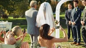Wedding guests raise their glasses in celebration during an outdoor garden ceremony near a white arch.