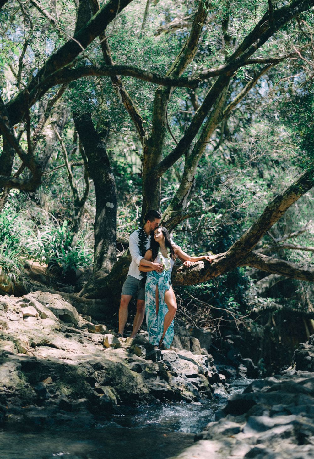 A couple stands together on rocks near a stream in a tropical forest setting.