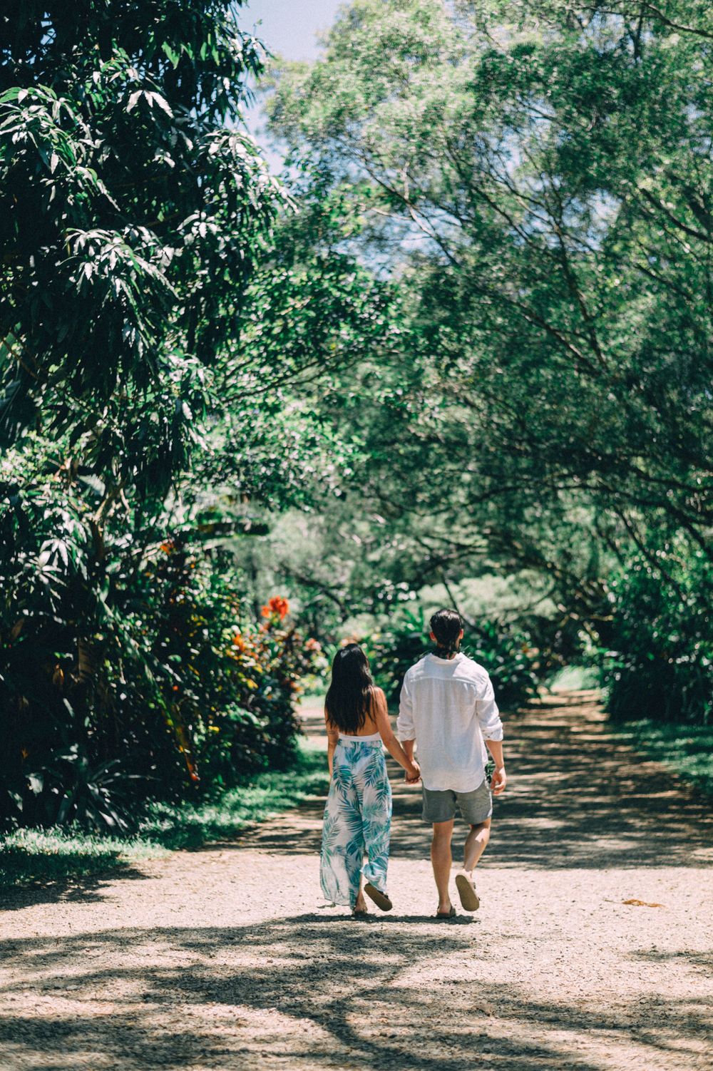 A couple walks hand in hand down a sun-dappled garden path surrounded by tropical trees.
