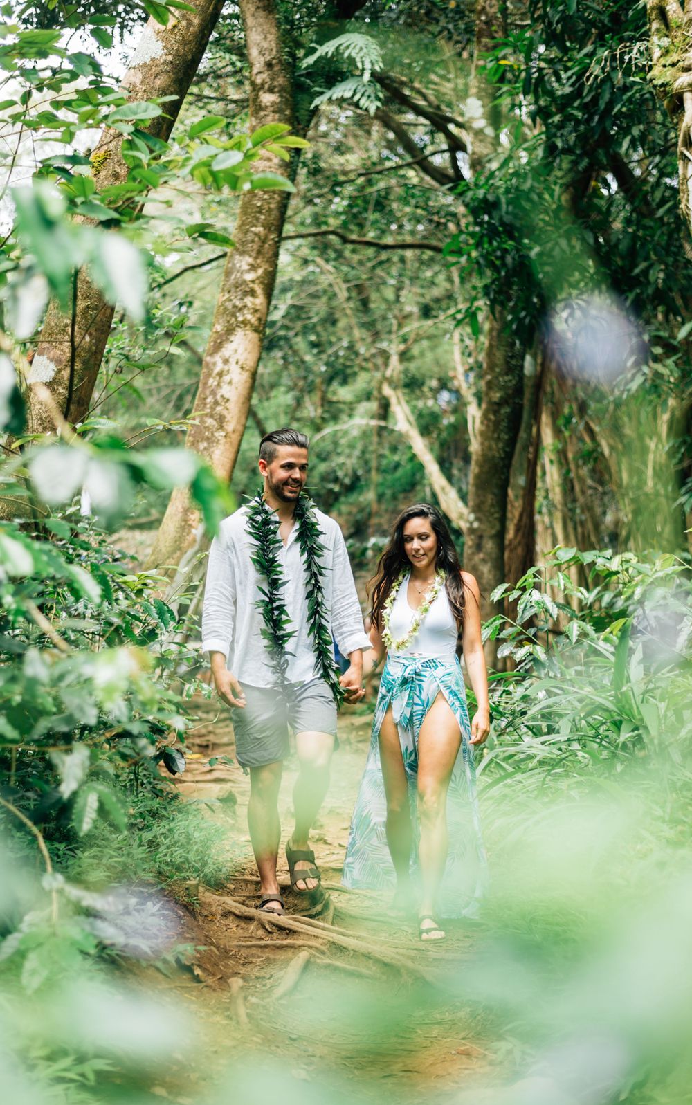 A couple walks hand in hand along a forest trail wearing traditional Hawaiian leis.