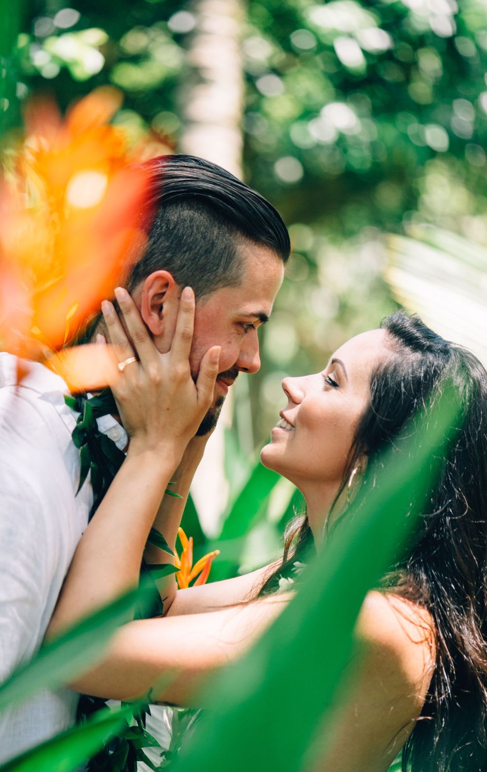 A romantic couple shares an intimate moment among tropical foliage with orange flowers in the background.