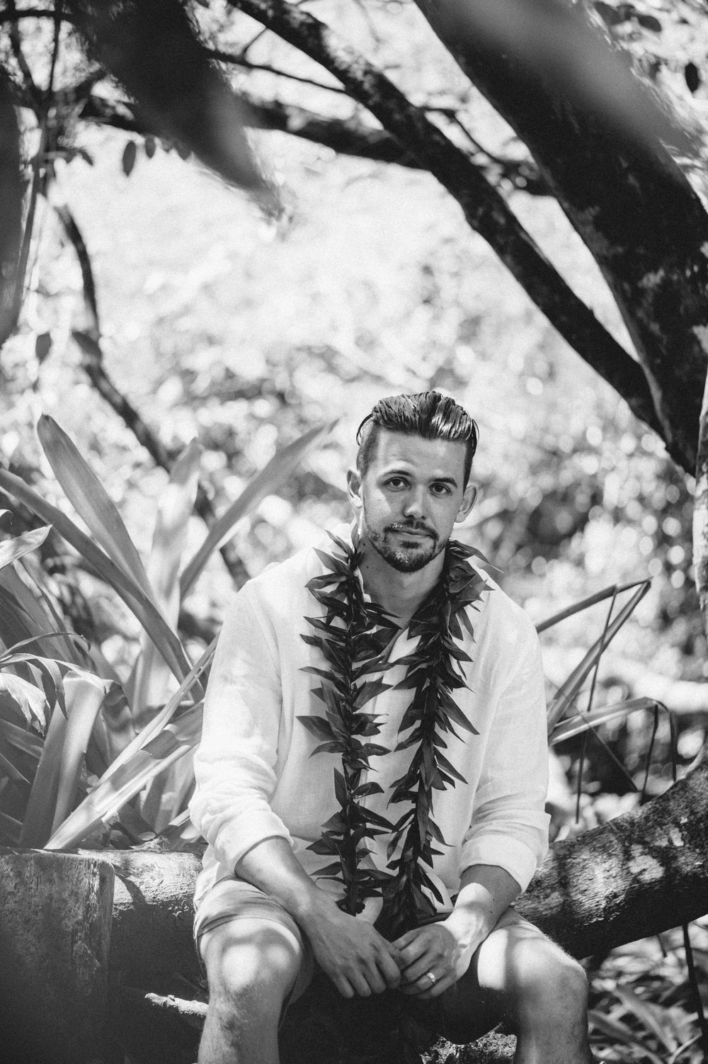 A person wearing a traditional black lei sits on a rock in a natural outdoor setting.