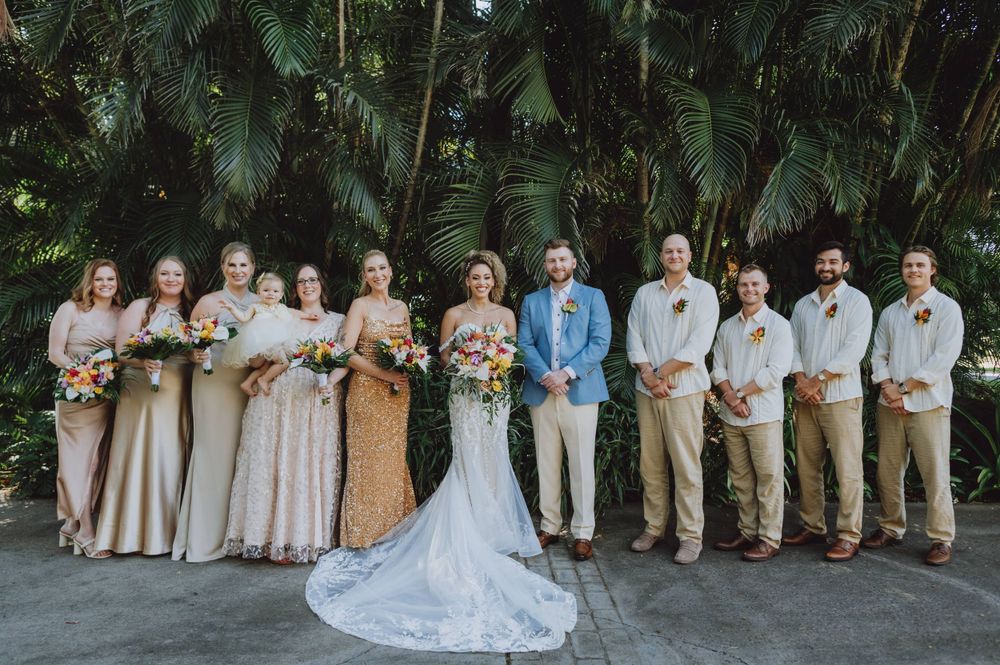 Wedding party poses for group photo against lush tropical garden backdrop.