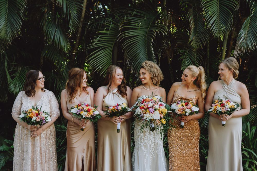 Bridesmaids in neutral dresses hold bouquets while standing in a tropical garden setting.