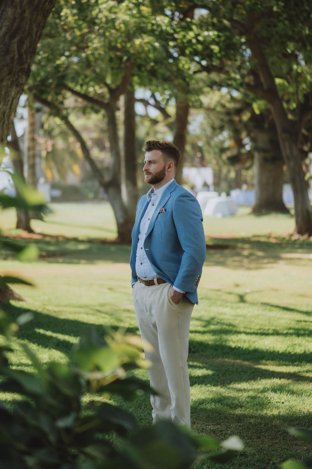 Groom in light blue suit walking through a sunlit garden setting.