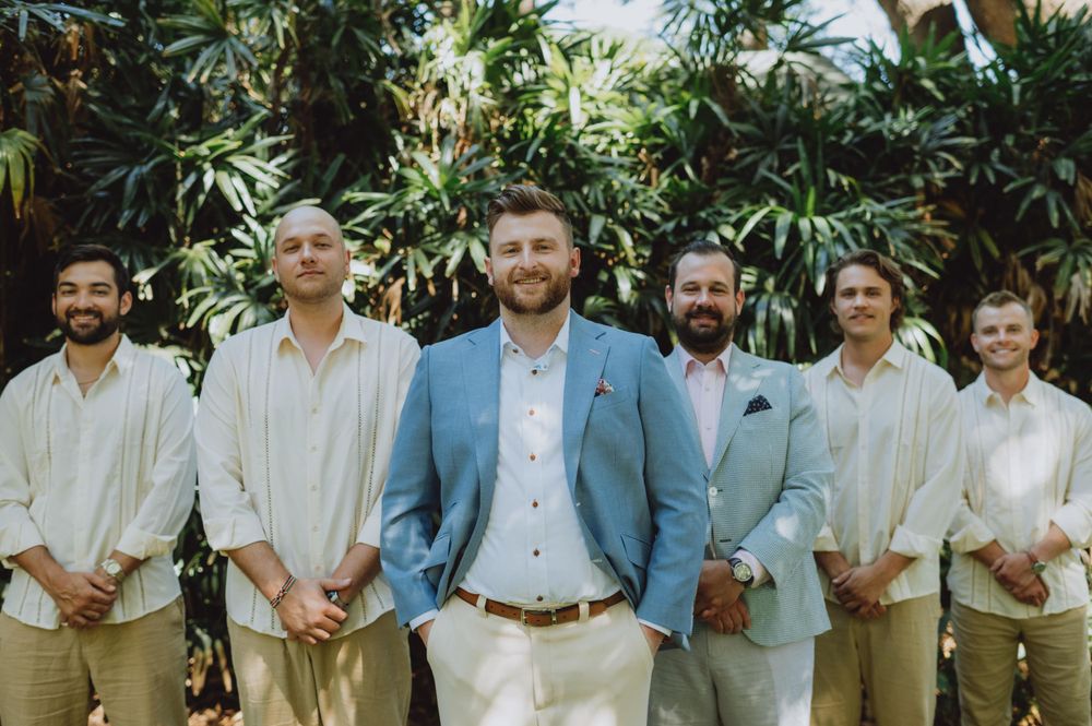 Groomsmen in light colored suits and blue shirts pose together in a tropical garden.