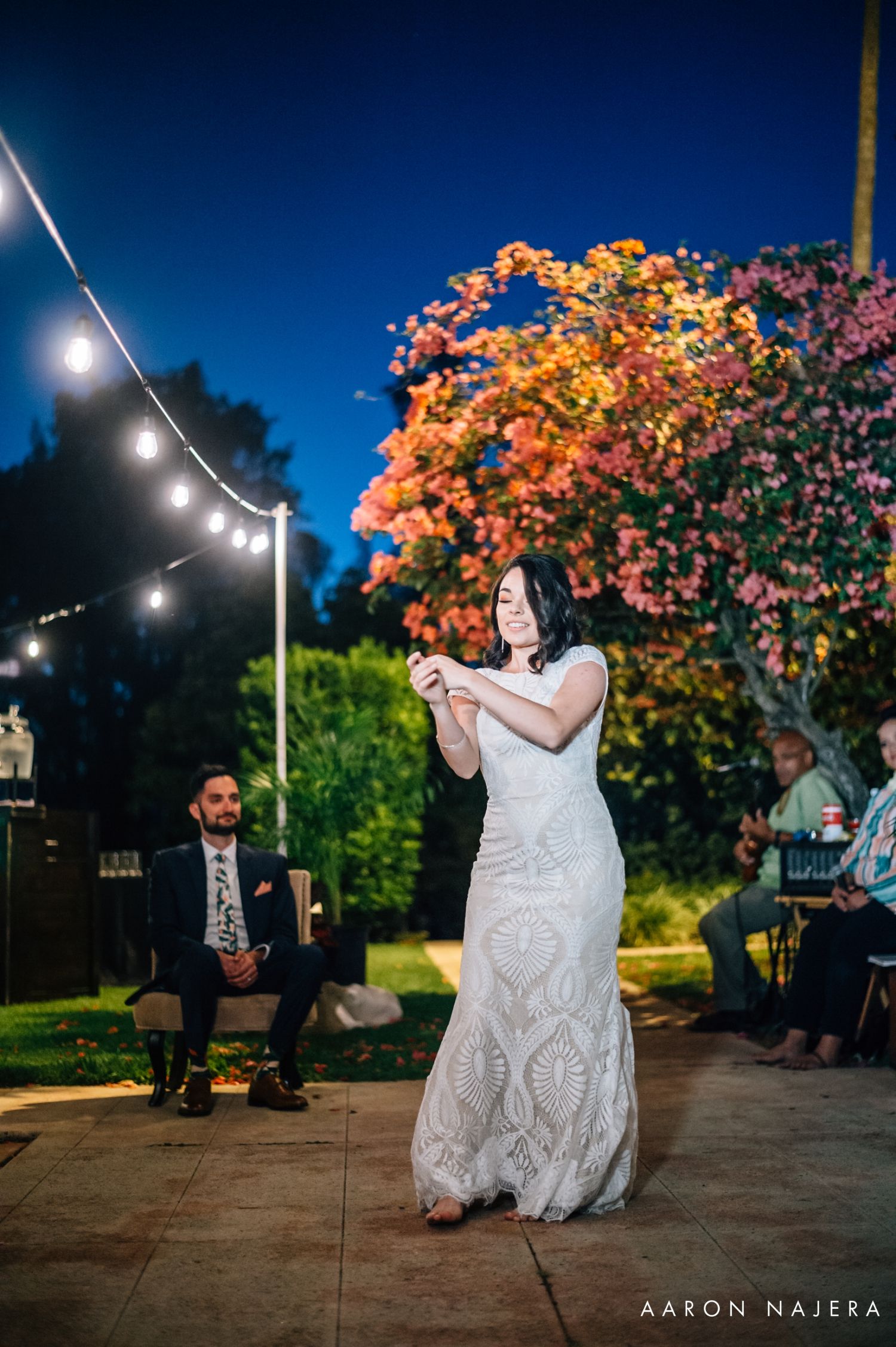 A couple dances at an outdoor evening wedding reception under string lights and colorful autumn trees.
