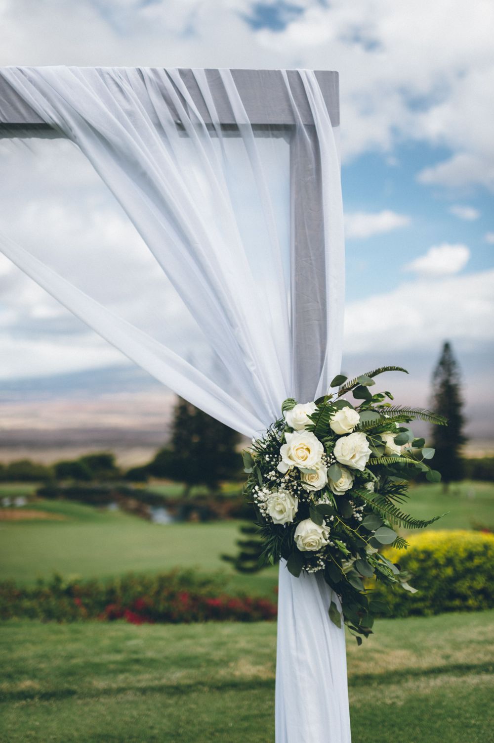 White wedding arch decorated with elegant white roses and greenery against a scenic mountain backdrop.
