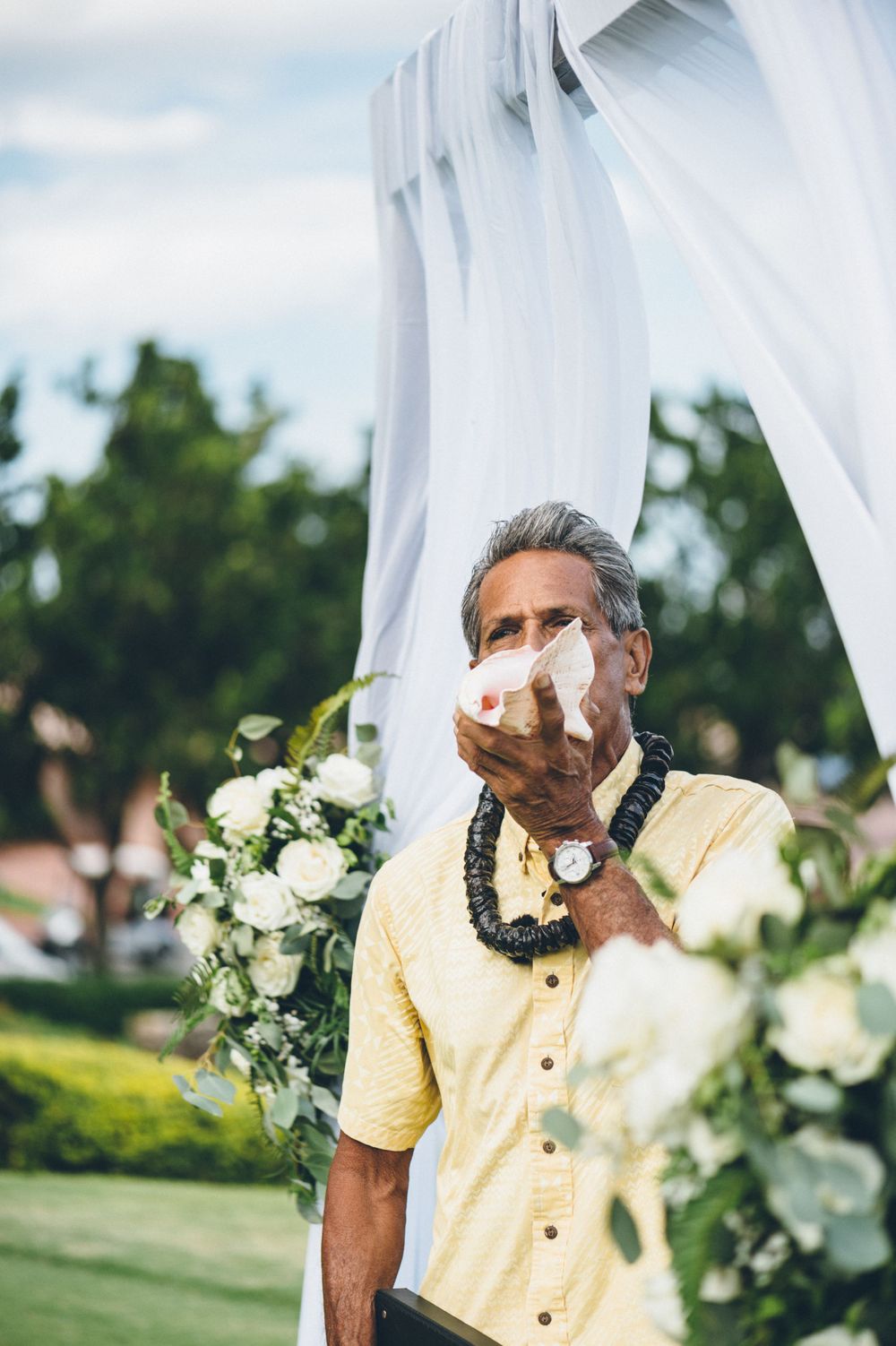 Person in yellow shirt with traditional lei blows into conch shell at outdoor wedding.