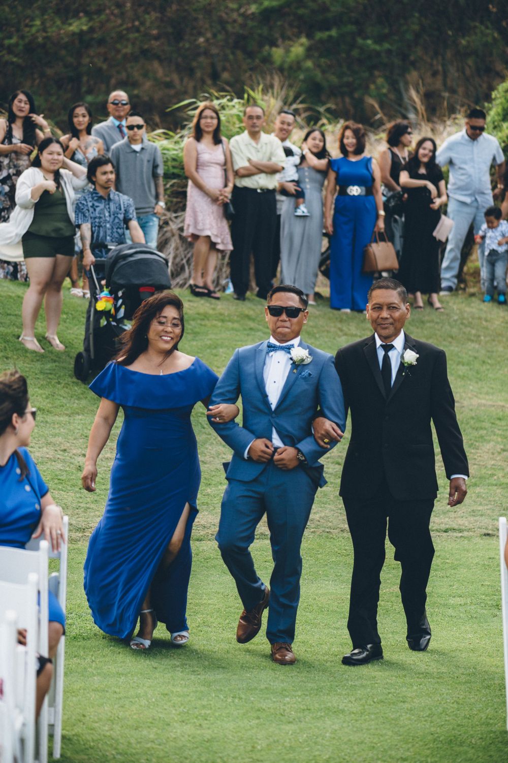 Wedding party members walk down a grassy aisle in coordinated blue and black formal attire.
