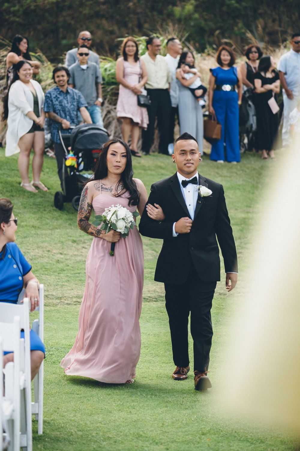Wedding party members walking down the aisle in formal attire on a grassy lawn.
