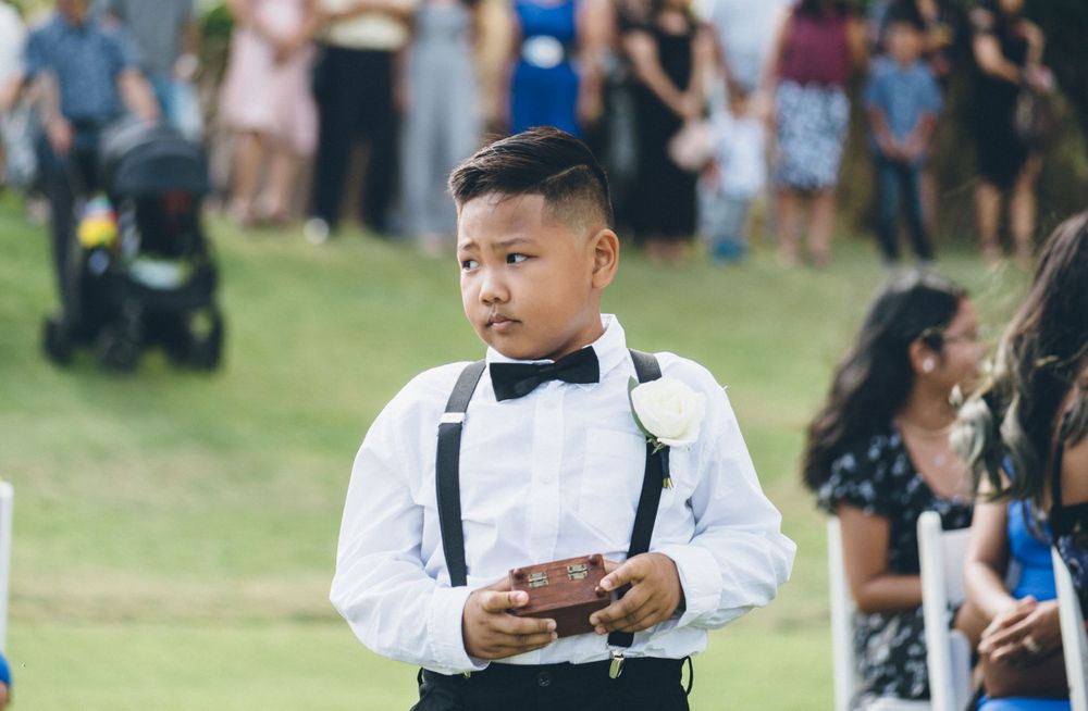 Young attendant in white shirt and bow tie stands formally at outdoor wedding ceremony.