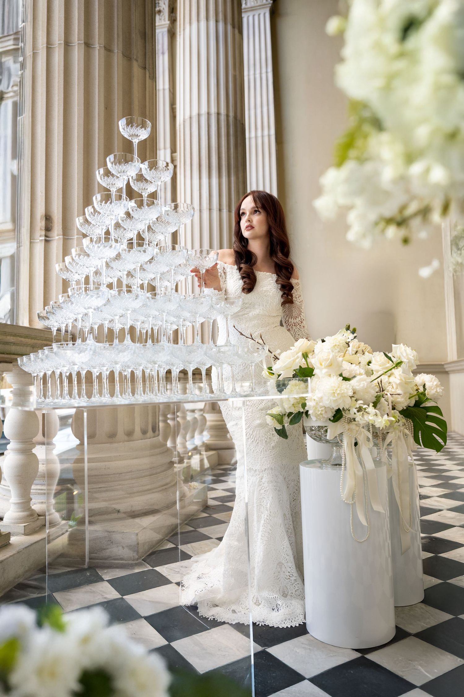 Elegant bride in lace gown standing beside a champagne tower and white floral arrangements in a grand hall.