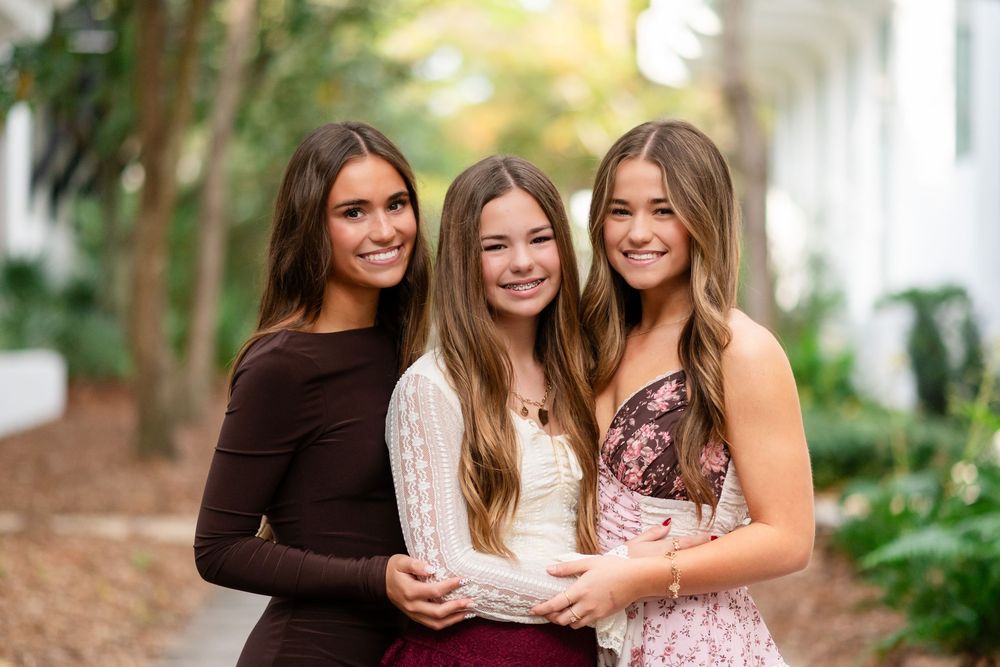 Three friends smile and laugh together while posing outdoors in casual attire during a sunny day.