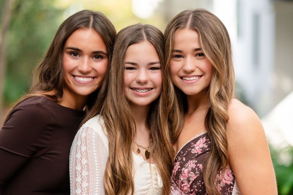 Three people smiling together outdoors in natural lighting with greenery in the background.