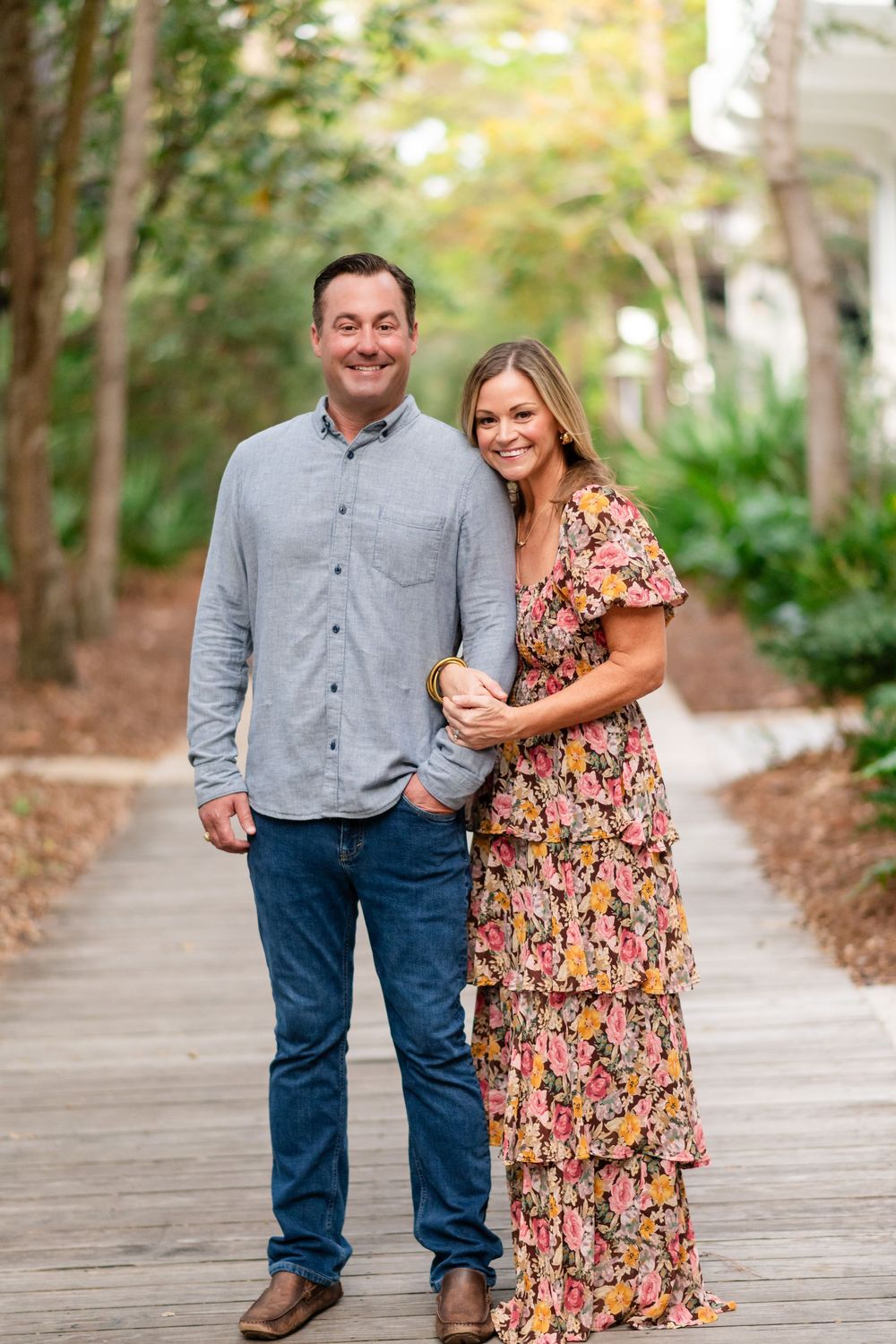 A couple poses together on a wooden boardwalk surrounded by trees and greenery in an outdoor setting.