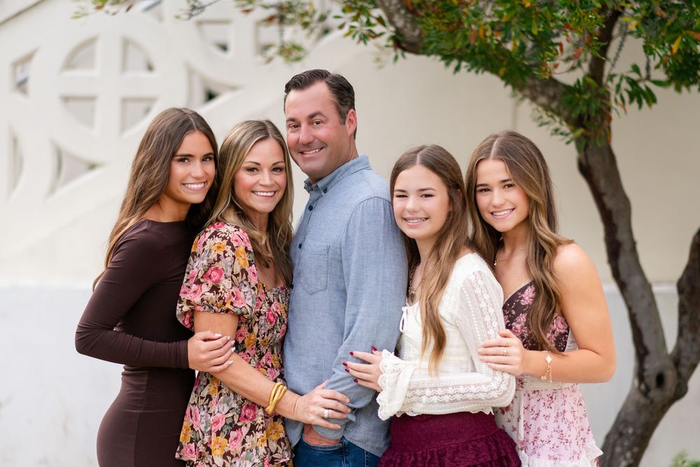 A family group poses together outdoors in coordinating outfits near a white building and tree.