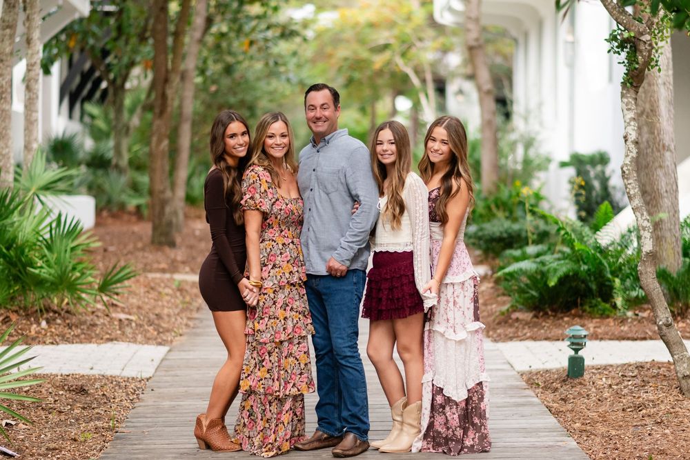A group of people walk and laugh together on a tree-lined path covered in pine needles.