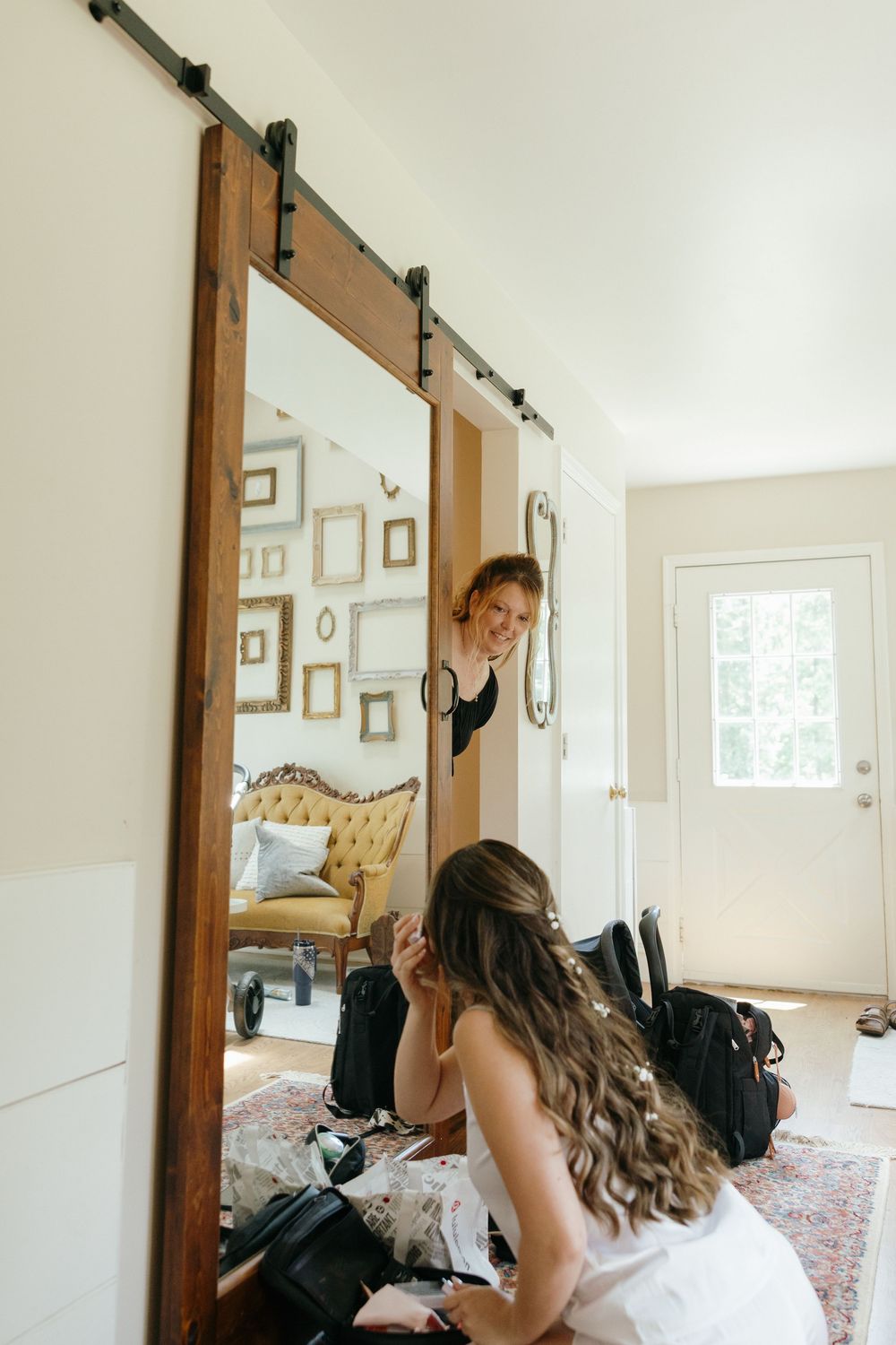 A sliding barn door mirror reflects people getting ready in a bright room with vintage furniture and framed wall art.