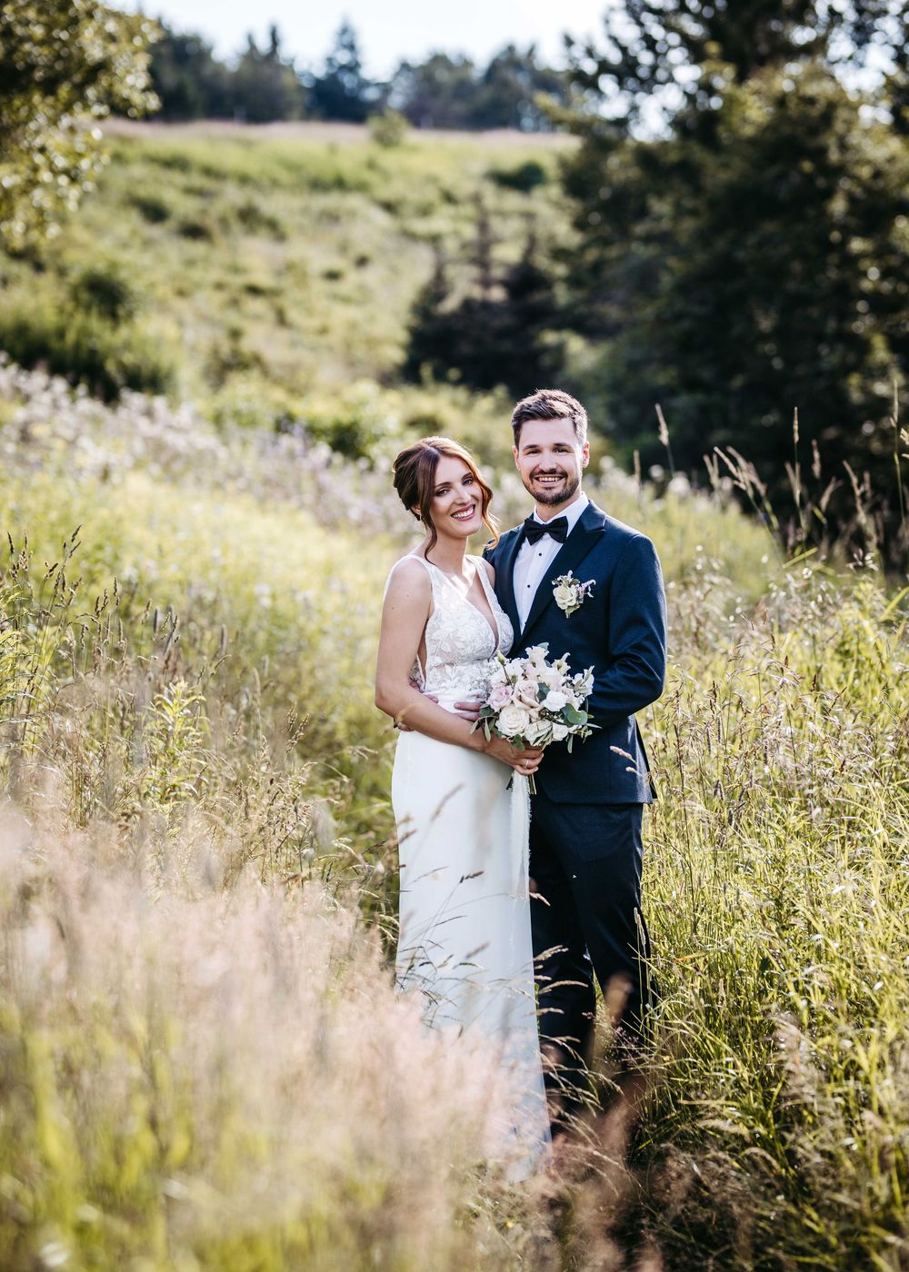 A couple stands together in a sun-drenched meadow filled with wildflowers and tall grass on their wedding day.