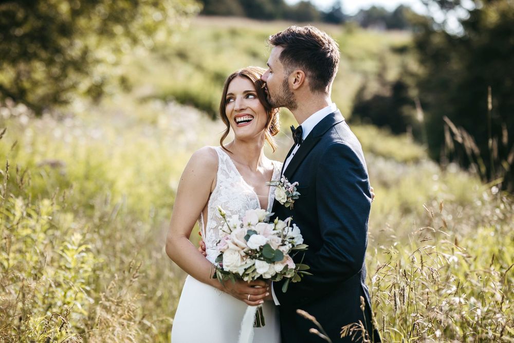 A romantic wedding couple embraces in a sunlit meadow with white floral bouquet and black suit.