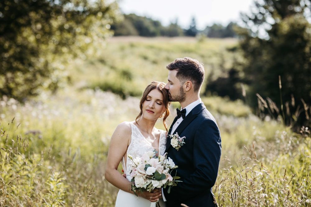 A couple in wedding attire embraces in a sunlit meadow with tall grass and trees in the background.