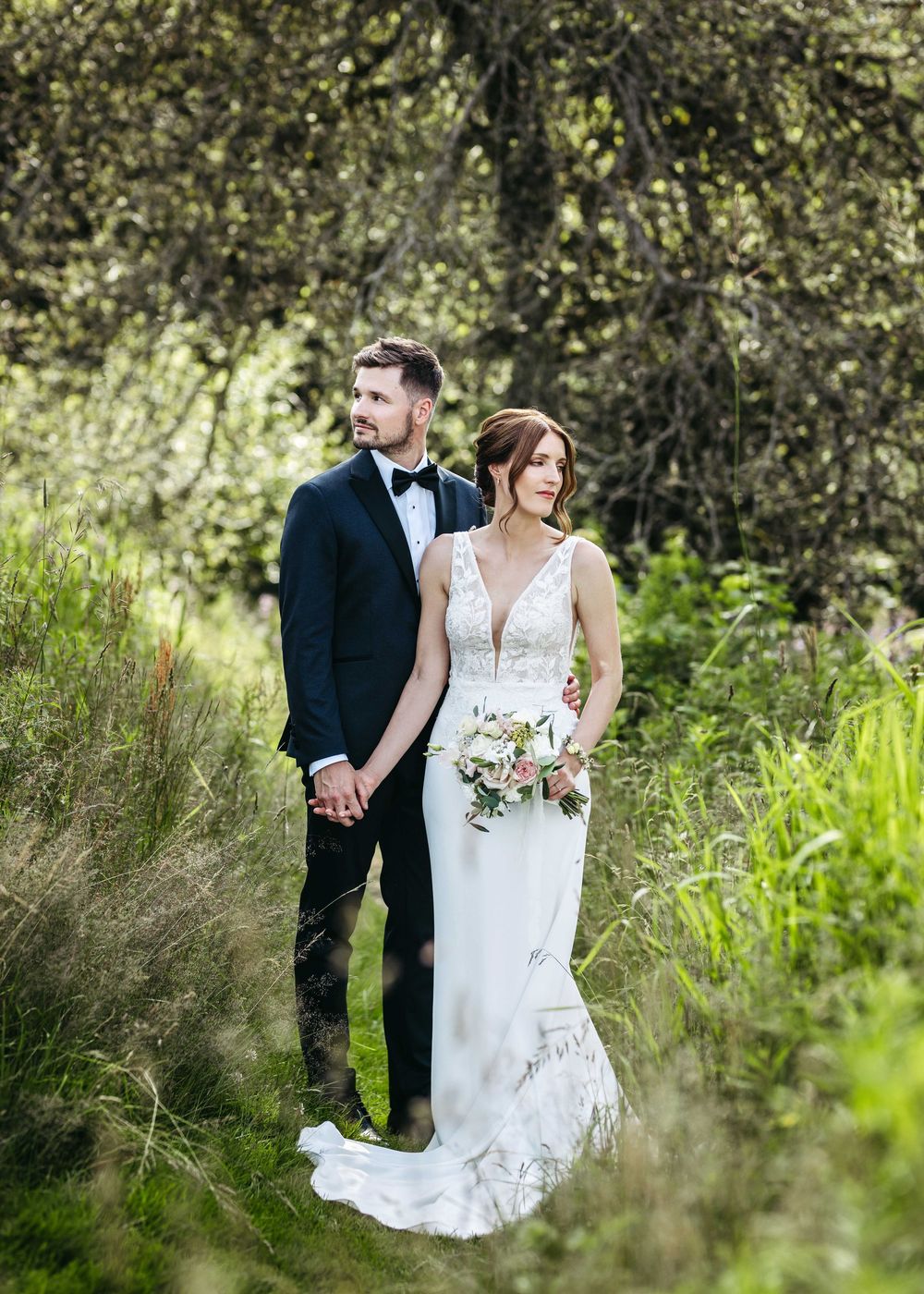 A couple in formal wedding attire pose together in a lush outdoor setting with natural greenery and sunlight.