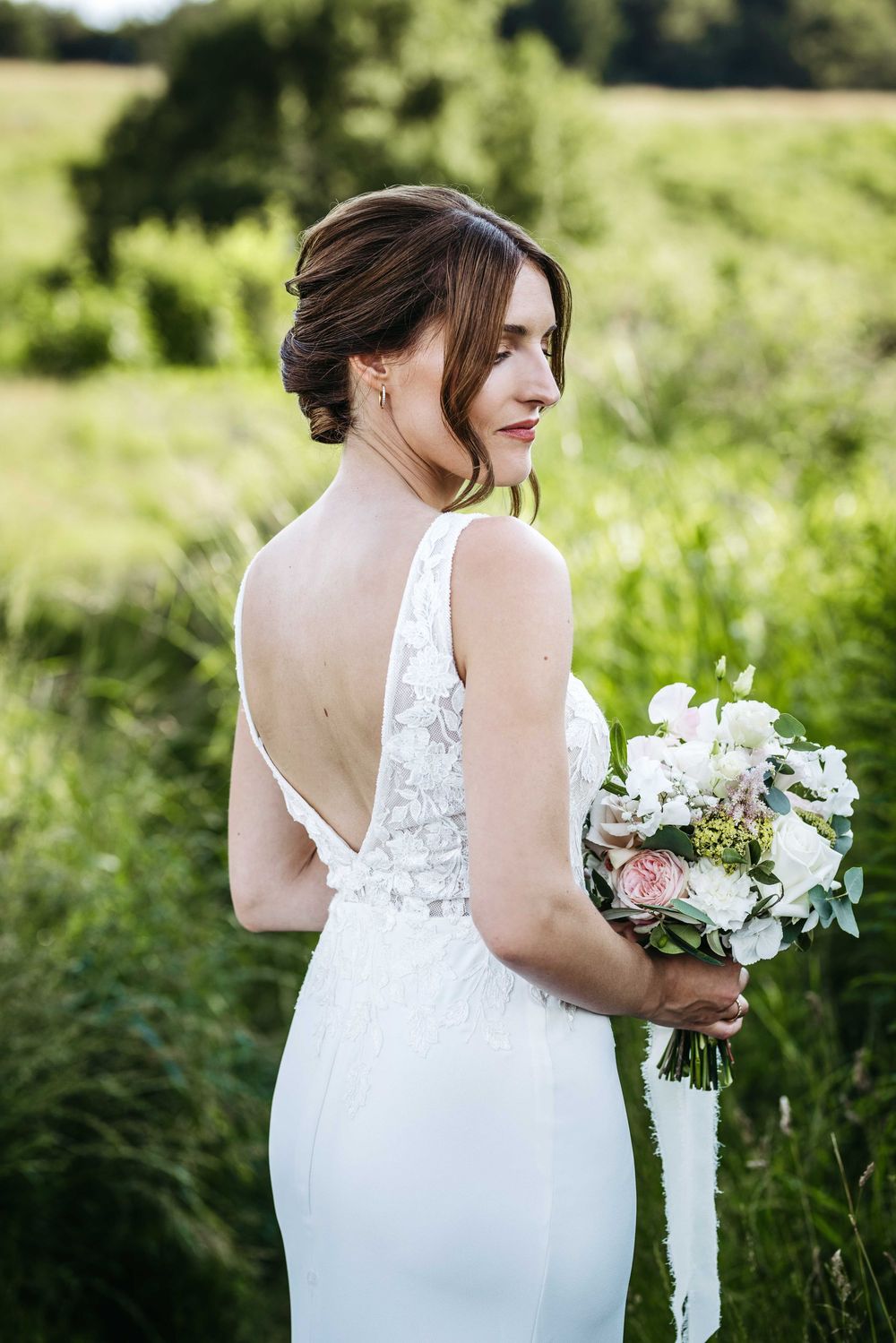 A bride in a white lace wedding dress with low back detail holds a bouquet of white and pink flowers in a green field.