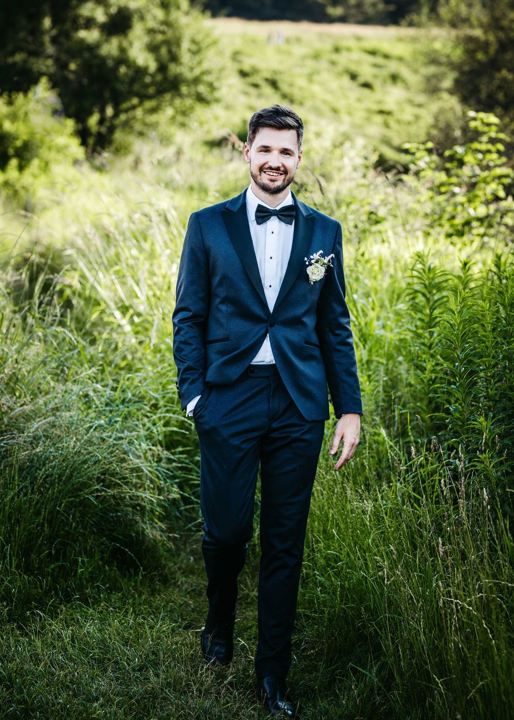 A groom in a classic black tuxedo and bowtie poses against lush green grass and foliage in a natural outdoor setting.