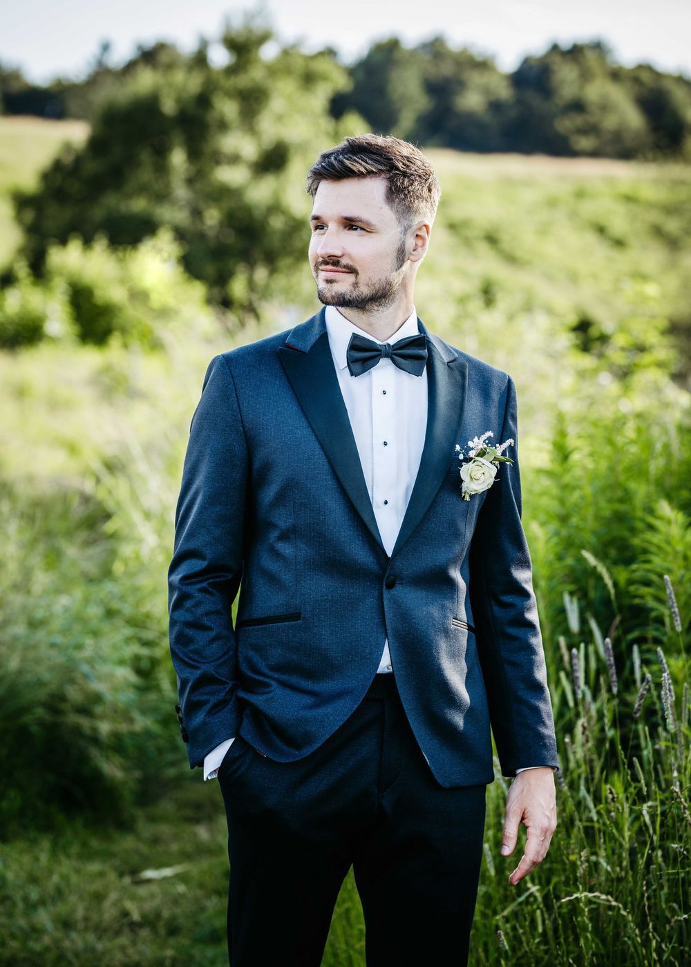 Groom in navy tuxedo and black bow tie poses in a sunny meadow with tall grass and natural lighting.