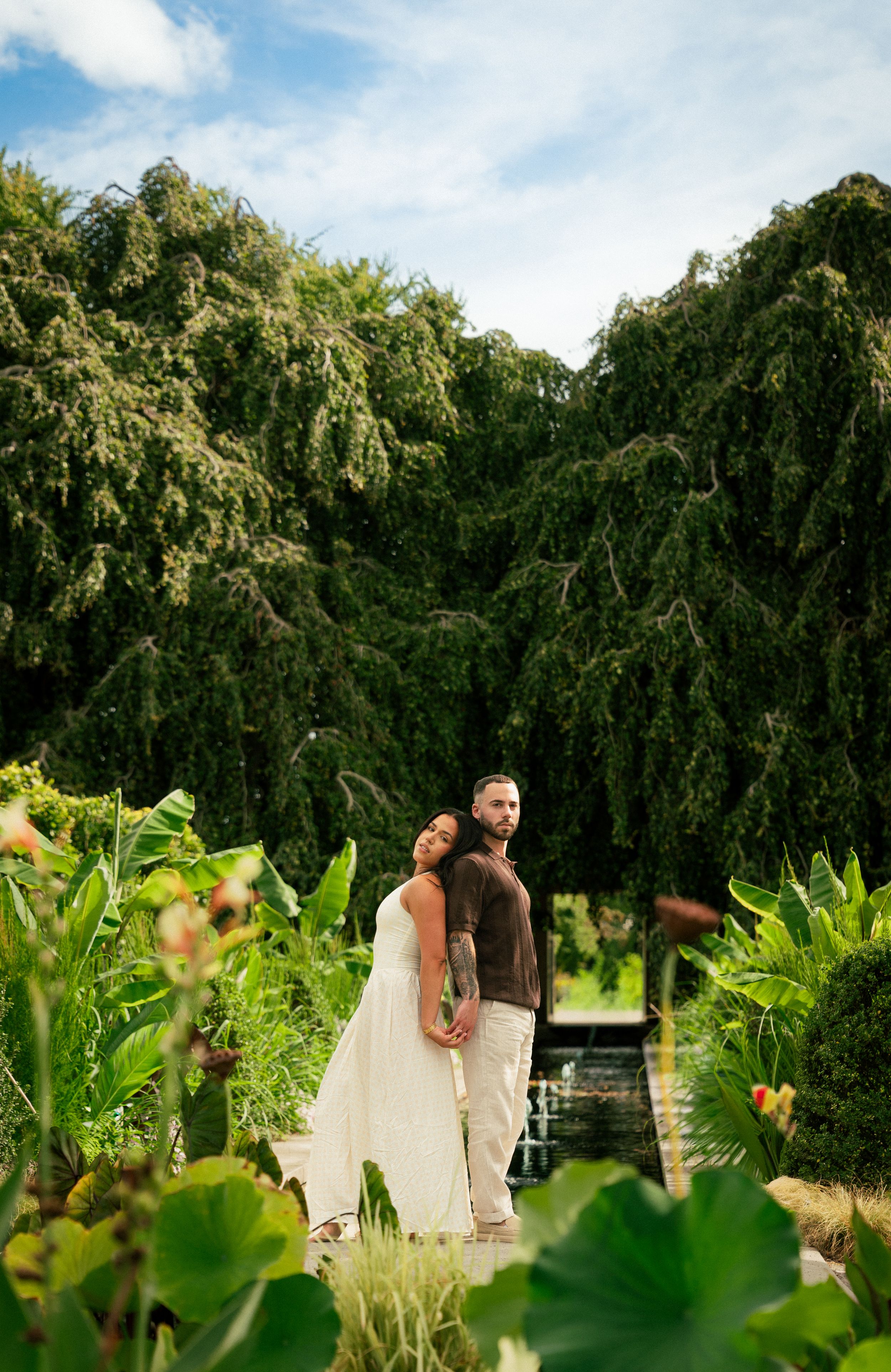 A couple shares an intimate moment among weeping willow trees and lush garden foliage.