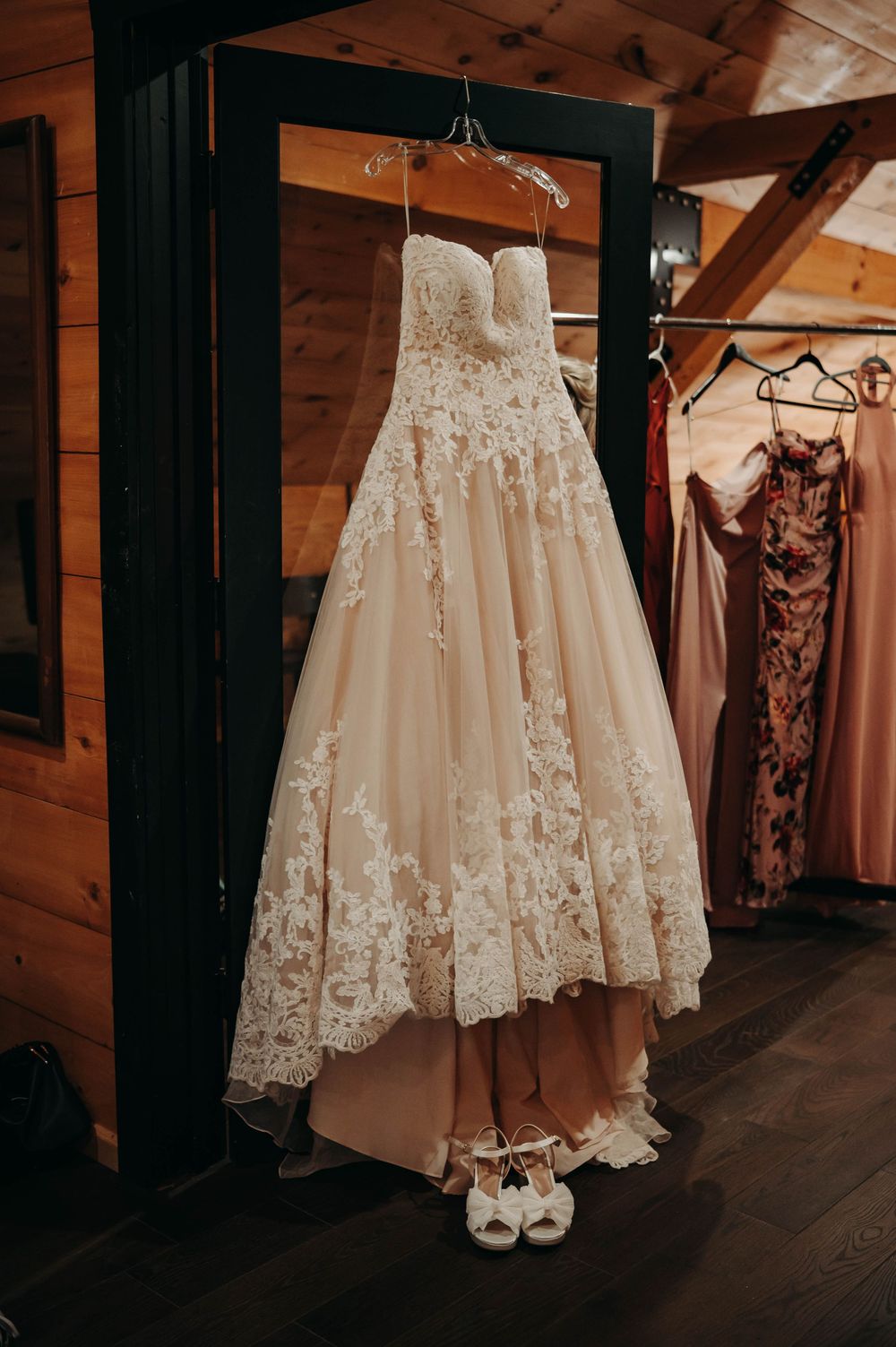 Elegant strapless lace wedding gown hanging in a rustic wooden bridal suite with bridesmaid dresses.