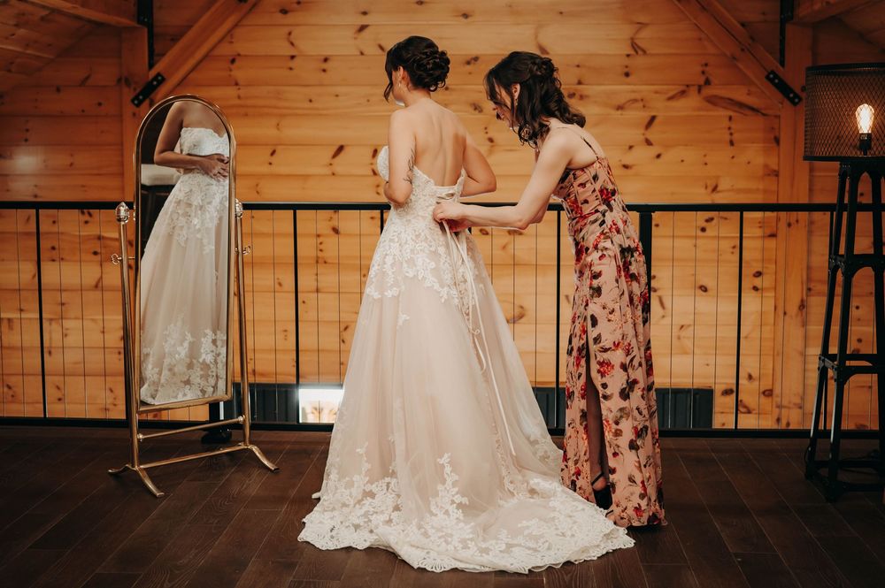 Bridesmaid in floral dress helping bride button her lace wedding gown in a rustic wooden bridal suite.