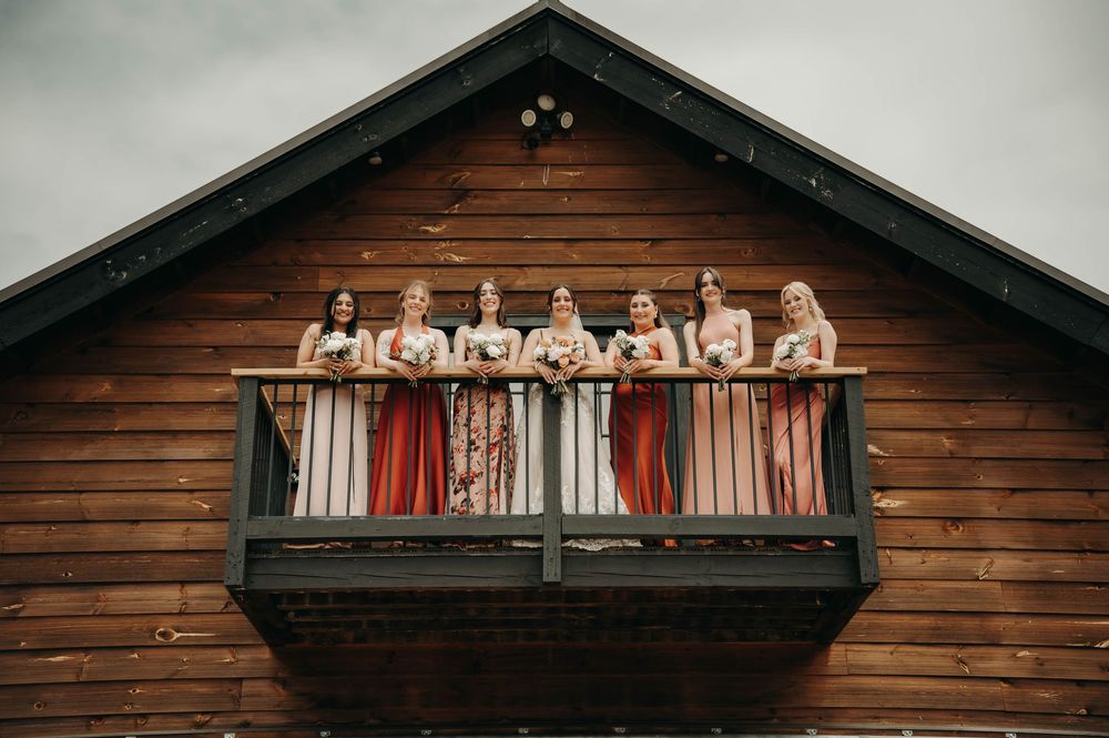 Bridal party in mismatched rust and blush bridesmaid dresses posing on a wooden barn balcony.