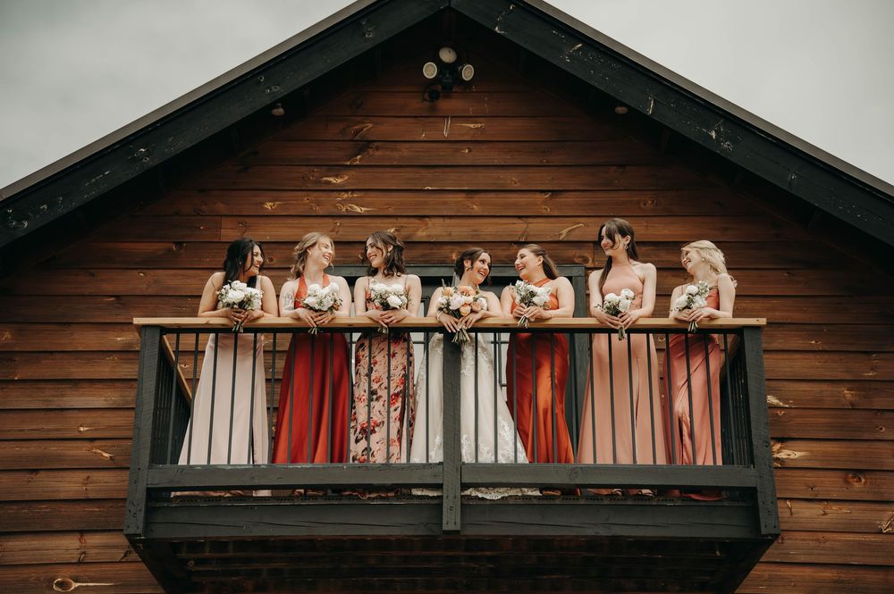 Bridesmaids in coordinating earth-tone dresses laughing together on a rustic barn balcony.